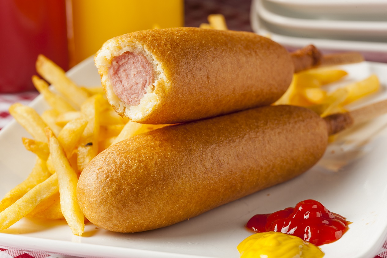 Zwei mit einem Teigmantel umhüllten Bockwürstchen auf einem Holzstiel. Ein Corn Dog ist im Anschnitt zu sehen.